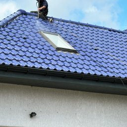 Roofers Folding Group - Czyszczenie niebieskiej dachówki ceramicznej myjką ciśnieniową, widoczny fragment okna dachowego i spływająca woda z rynny.