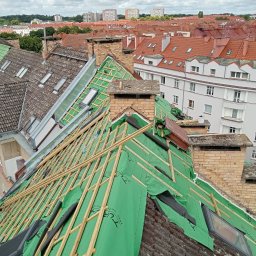Roofers Folding Group - Dach w trakcie remontu, widoczne drewniane łaty, zielona membrana dachowa, kominy z cegły, okna dachowe i panorama miasta w tle.