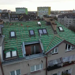 Widok z góry na dach budynku w trakcie przygotowania do nowego pokrycia, widoczna zielona membrana dachowa, łaty drewniane, okna dachowe i fragmenty starego pokrycia, w tle zabudowa miejska.