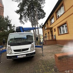 Roofers Folding Group - Praca na wysokościach: podnośnik koszowy przy budynku z żółtą elewacją, widoczny pracownik w koszu. W tle ceglany kościół i drzewa.