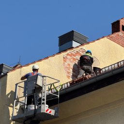 Roofers Folding Group - Dwóch pracowników na podnośniku naprawia uszkodzony fragment elewacji budynku z ceglanym kominem i metalowymi wylotami wentylacyjnymi na dachu.