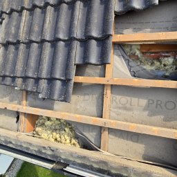 Roofers Folding Group - Fragment dachu w trakcie naprawy: czarne dachówki, drewniana konstrukcja, izolacja z wełny mineralnej i membrana dachowa z widocznym napisem 'DIVOROLL PRO'.