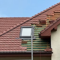 Roofers Folding Group - Częściowo zdjęte czerwone dachówki odsłaniające drewnianą konstrukcję dachu z oknem dachowym. Widoczny fragment elewacji budynku i rusztowanie.