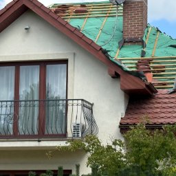 Roofers Folding Group - Dom z częściowo zdjętym dachem, widoczne drewniane łaty i zielona membrana. Fragment dachu pokryty czerwoną dachówką. Balkon z ozdobną balustradą.