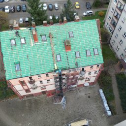 Roofers Folding Group - Widok z góry na budynek z zielonym dachem w trakcie krycia. Widoczne rusztowania, pracownicy i elementy konstrukcyjne dachu. W tle zaparkowane samochody i budynki.