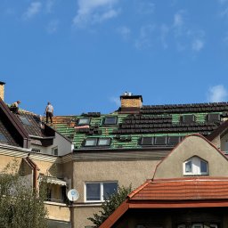 Roofers Folding Group - Fragment dachu z częściowo ułożoną dachówką, widoczne okna dachowe i dwóch dekarzy podczas pracy na tle błękitnego nieba z delikatnymi chmurami.
