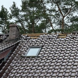 Roofers Folding Group - Częściowo zdjęte brązowe dachówki na dachu z oknem połaciowym i kominem z cegły. Widoczne elementy konstrukcji dachu. W tle drzewa iglaste.