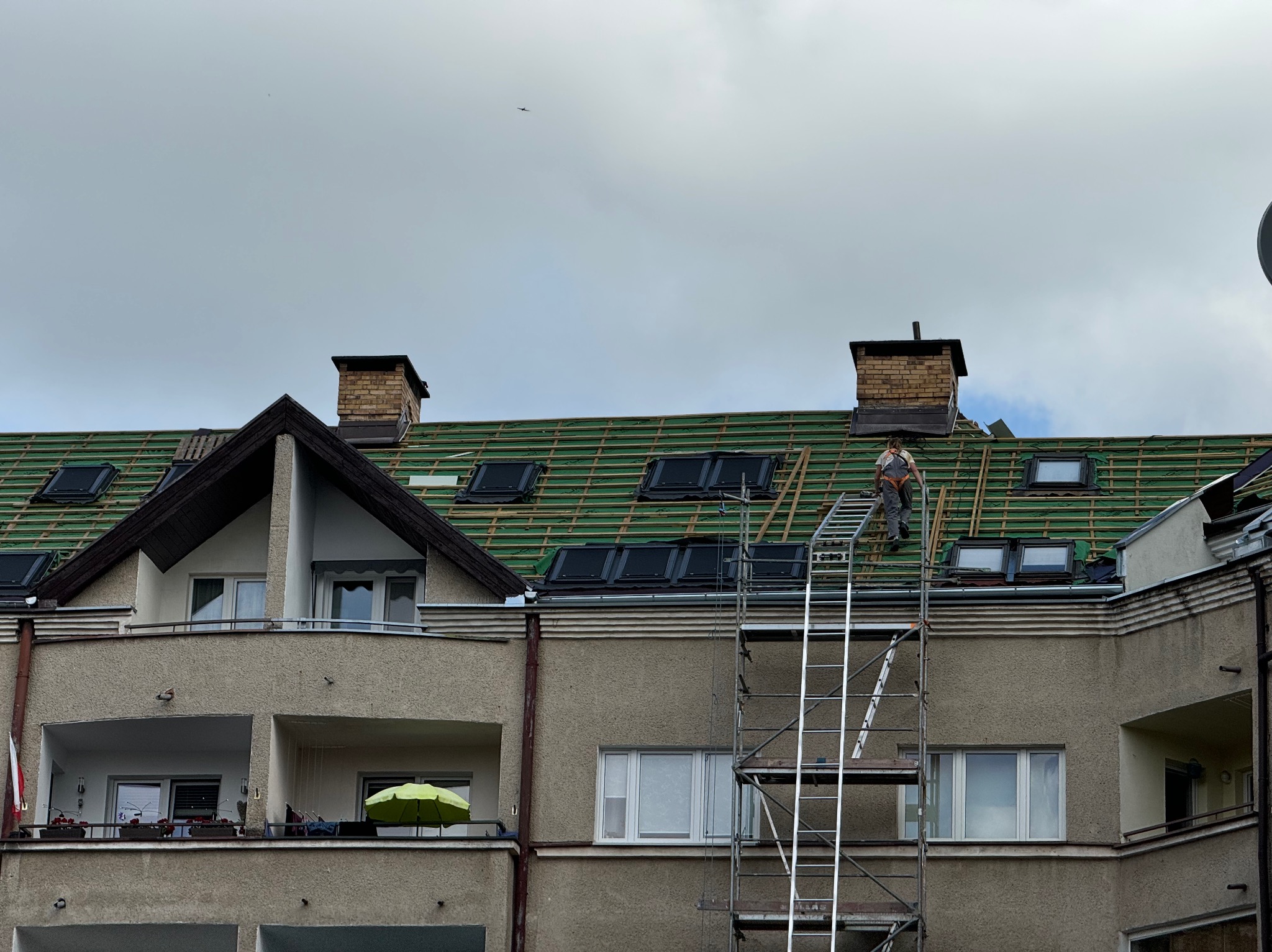 Pracownik na rusztowaniu montuje nowe okna dachowe na budynku z zieloną membraną dachową, widoczne kominy z cegły i przelatujący ptak na pochmurnym niebie.