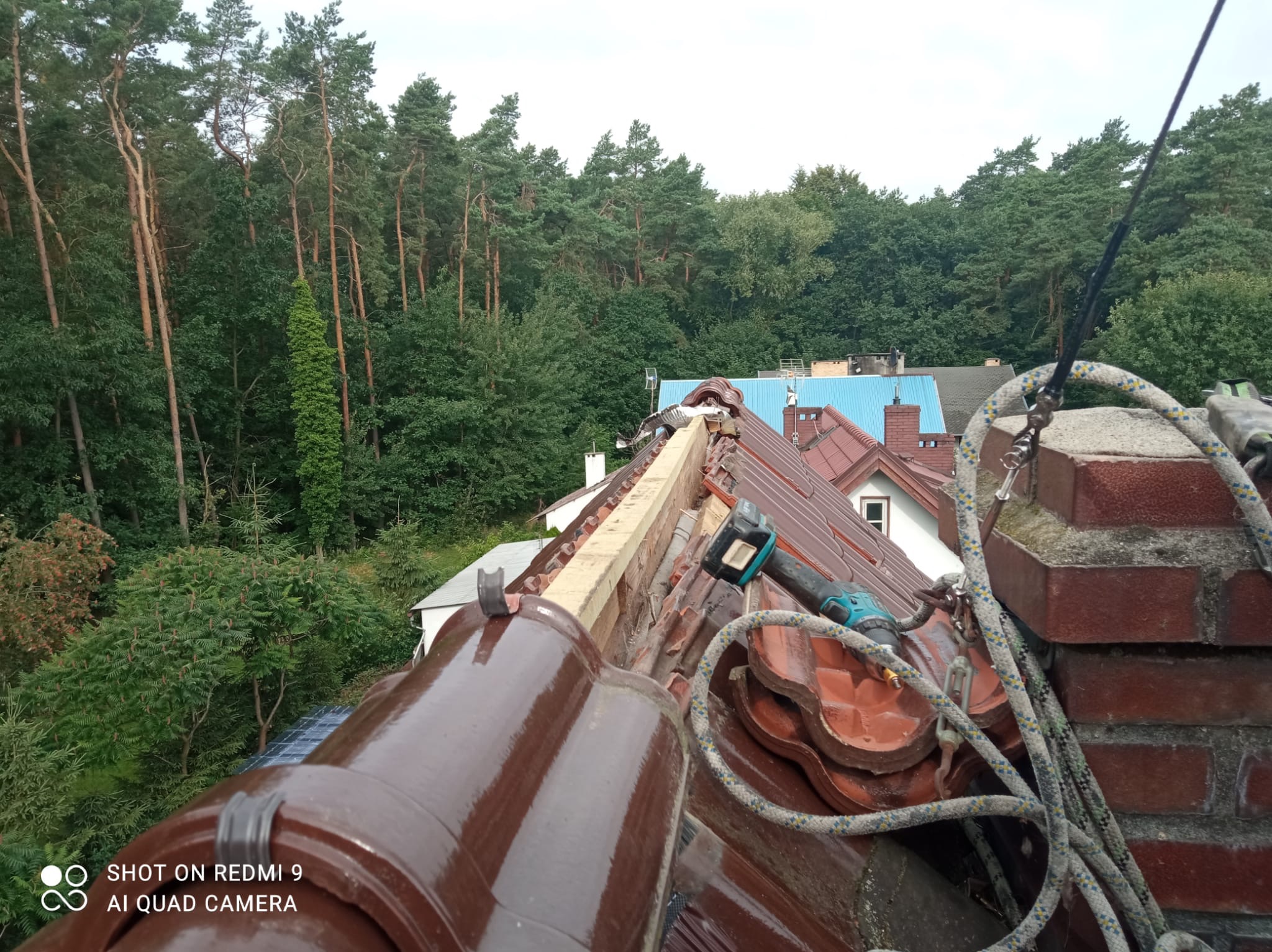 Fragment dachu z brązowej dachówki ceramicznej w trakcie prac dekarskich, widoczna wiertarko-wkrętarka akumulatorowa, liny zabezpieczające i drewniana łata, w tle zieleń drzew i inne dachy.