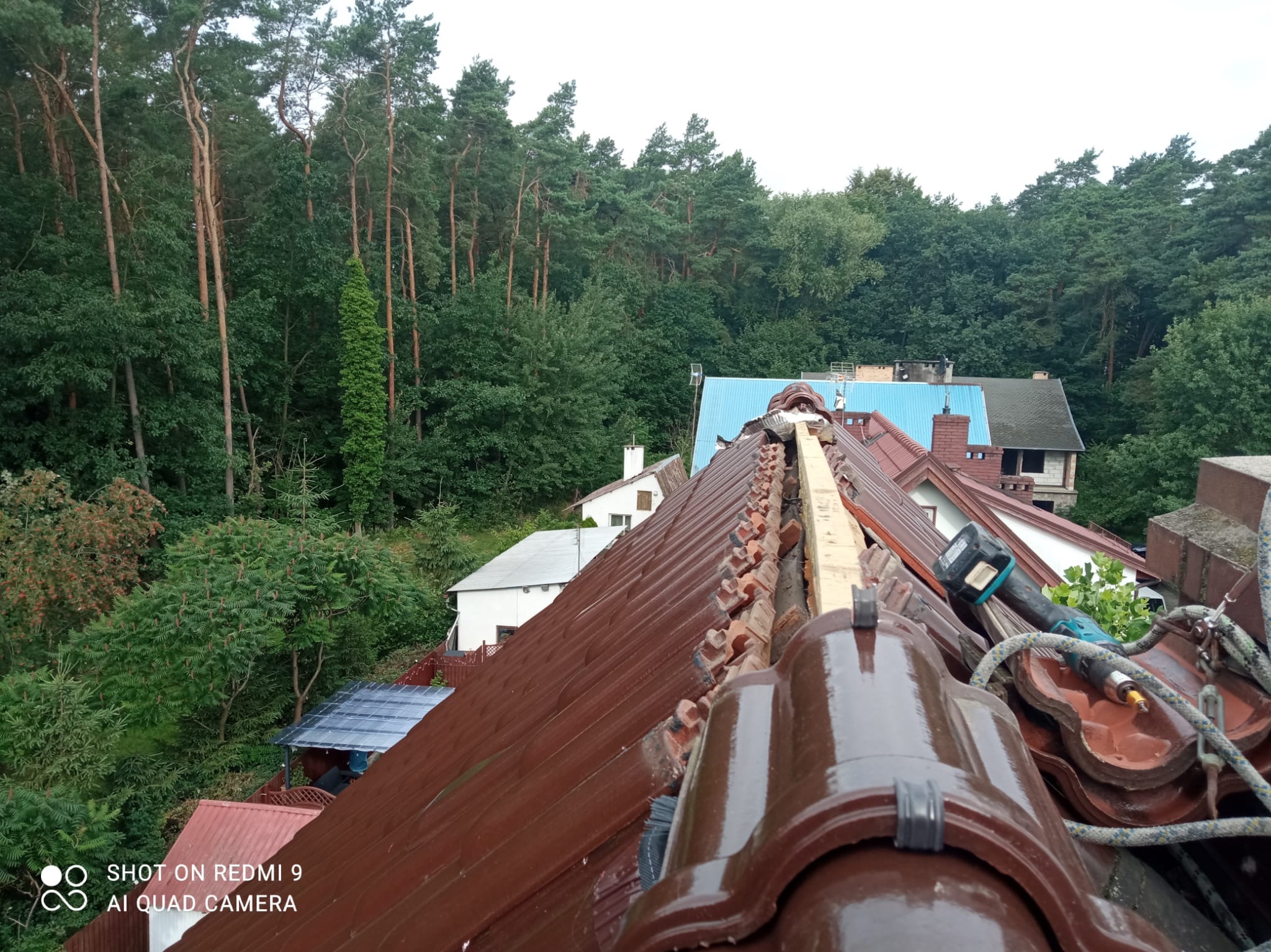 Brązowe dachówki ceramiczne podczas prac na dachu, widoczne narzędzia dekarskie, w tle las i zabudowania.