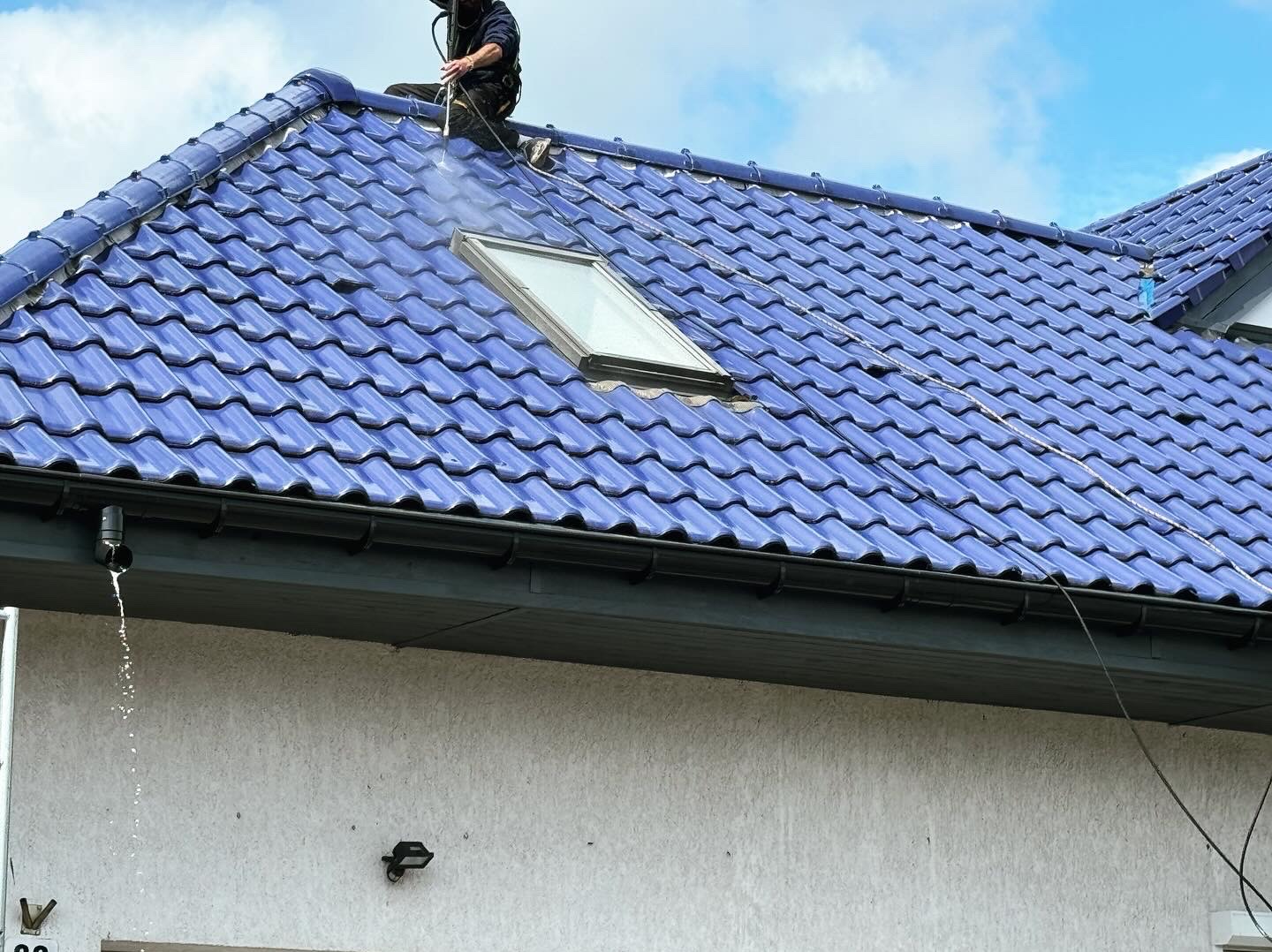 Czyszczenie niebieskiej dachówki ceramicznej myjką ciśnieniową, widoczny fragment okna dachowego i spływająca woda z rynny.