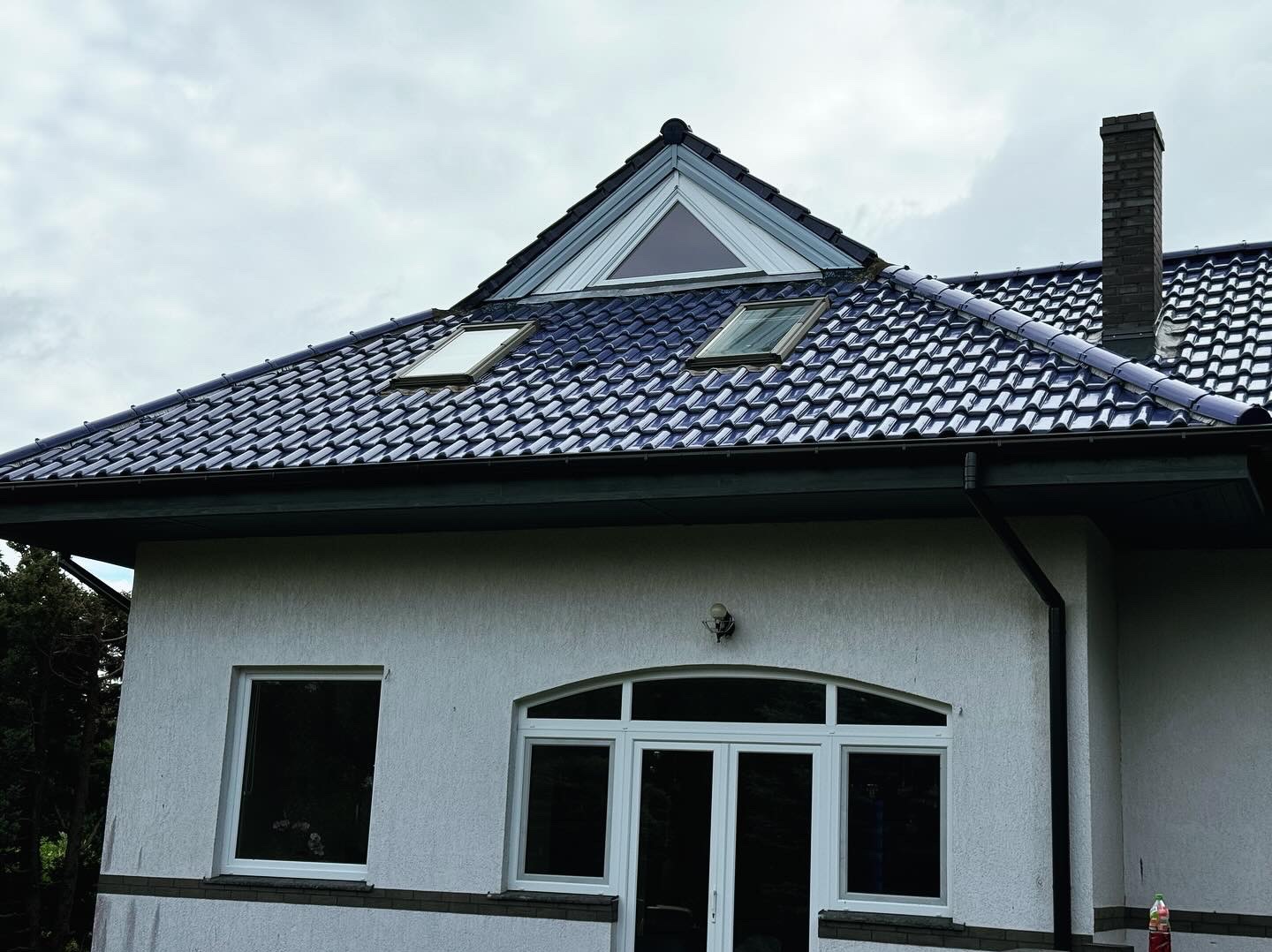 Dom z ciemną dachówką ceramiczną, dwoma oknami dachowymi i trójkątnym lukarną, widoczny komin z cegły i orynnowanie.