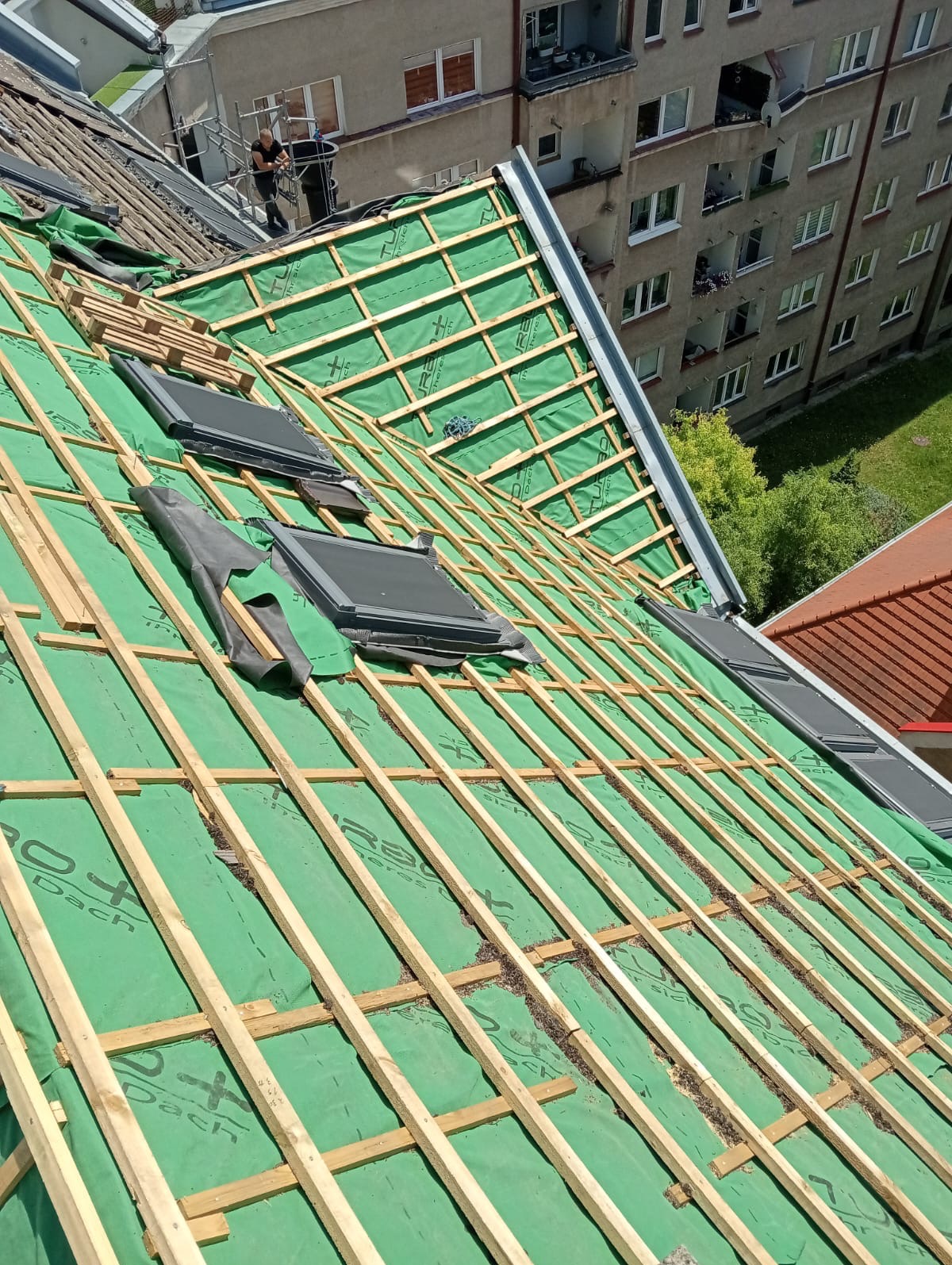 Montaż okien dachowych na konstrukcji z łat i kontrłat, pokrytej zieloną membraną, w tle budynek mieszkalny.