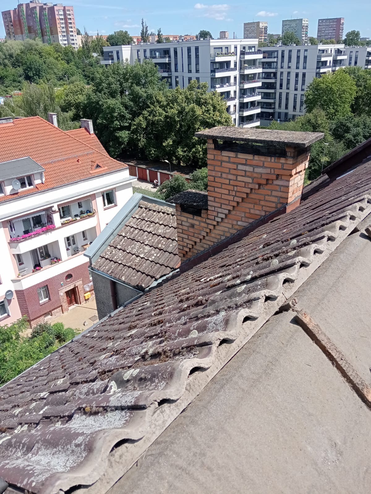 Zabrudzony dach z dachówki ceramicznej z mchem i porostami, widok z góry na komin z cegły i budynki mieszkalne w tle.