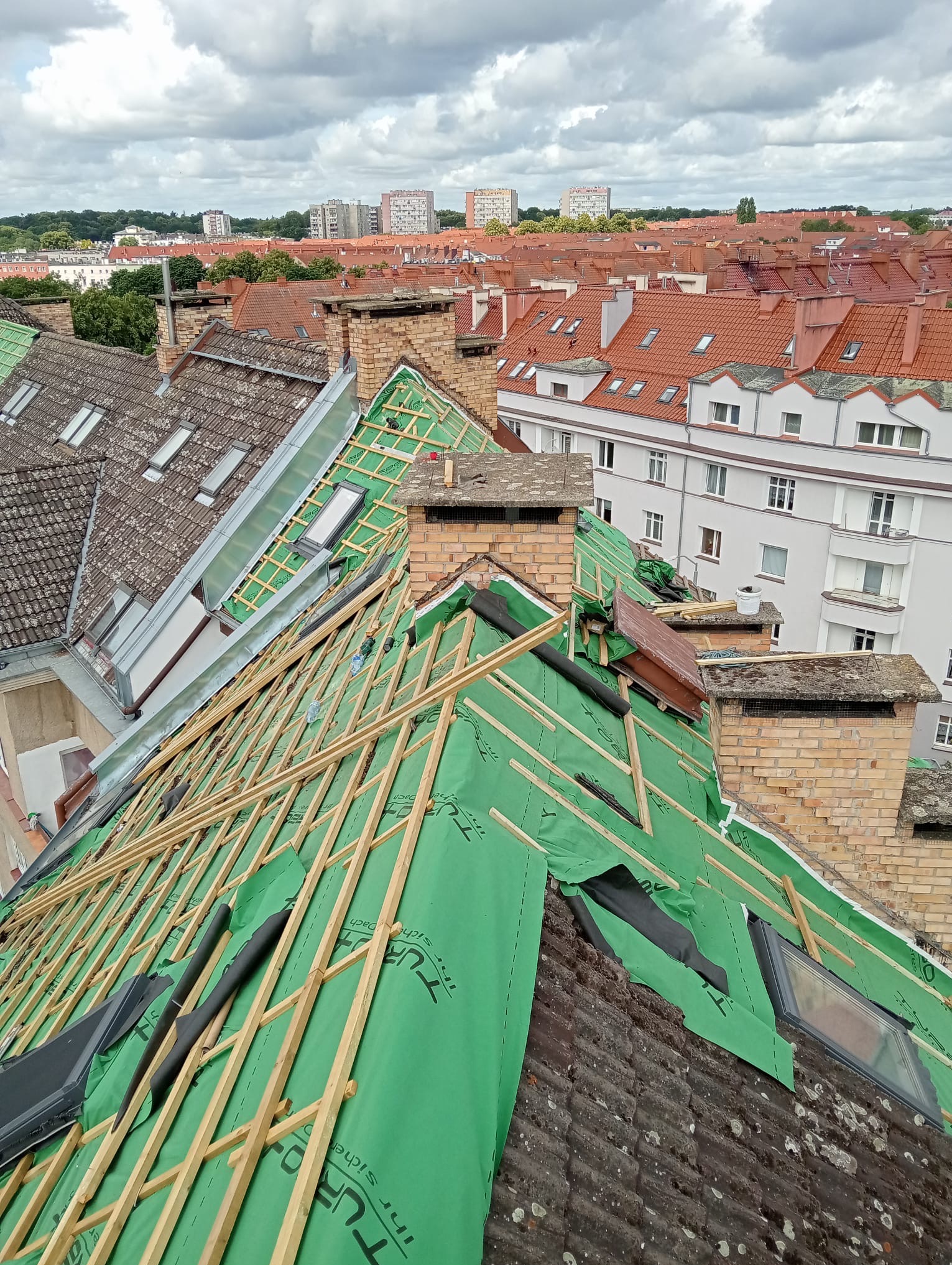 Dach w trakcie remontu, widoczne drewniane łaty, zielona membrana dachowa, kominy z cegły, okna dachowe i panorama miasta w tle.
