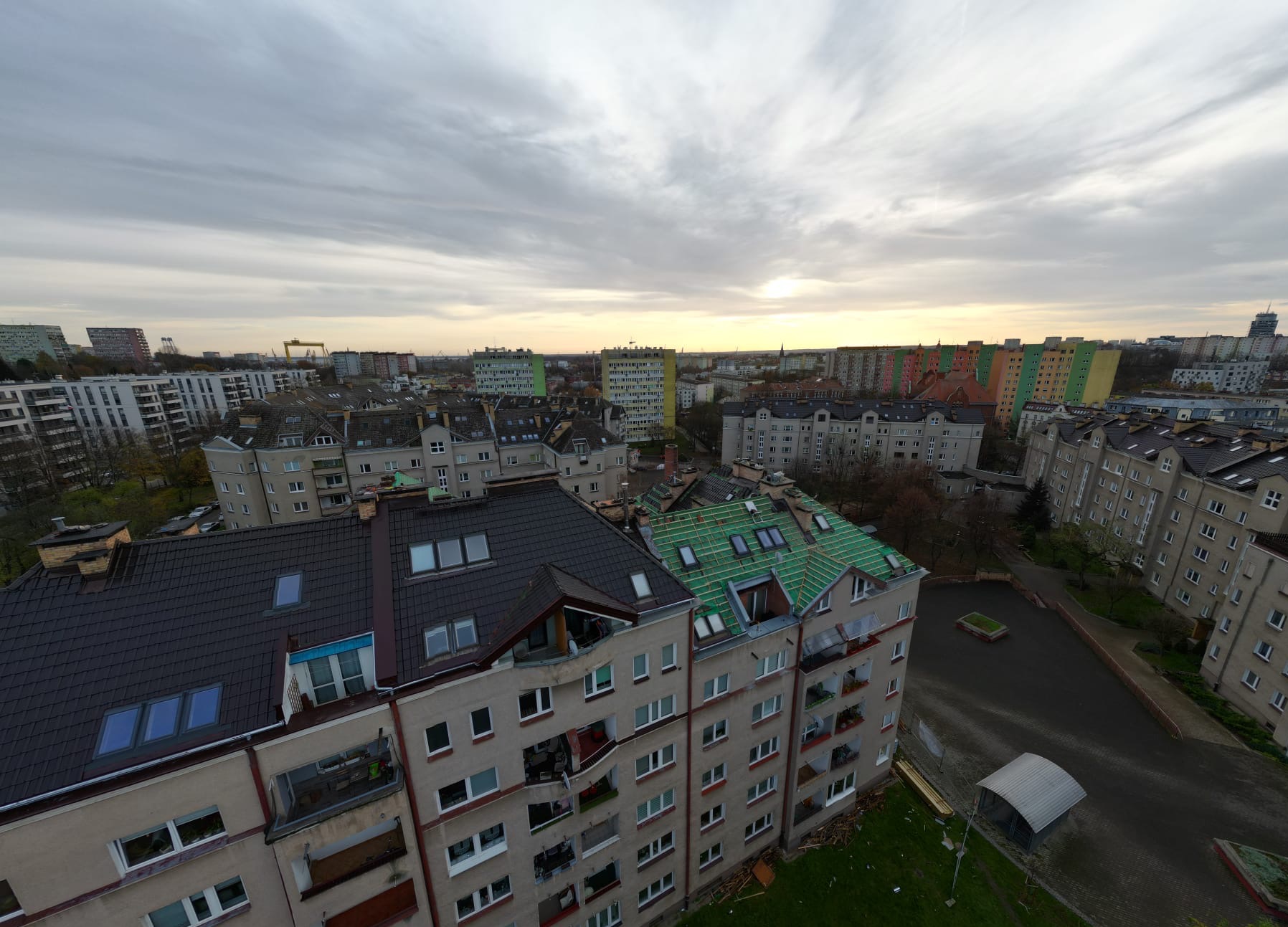 Panorama miasta z widokiem na budynek w trakcie prac dekarskich, gdzie widoczne są krokwie i zielona membrana na dachu, a w tle inne budynki i pochmurne niebo.
