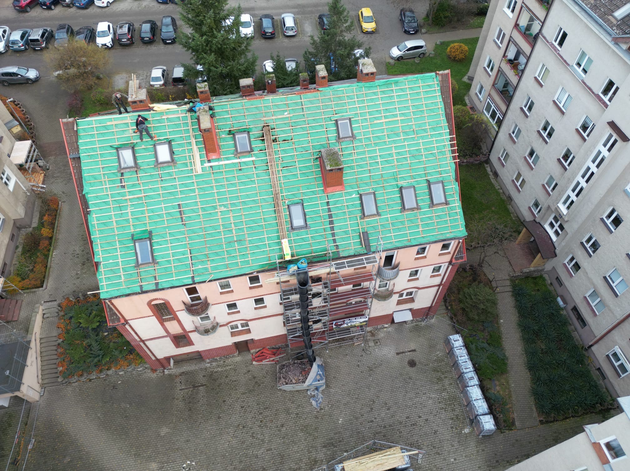 Widok z góry na budynek mieszkalny z demontowanym starym pokryciem dachu, widoczne rusztowania, ekipa pracowników, zielona membrana dachowa i przygotowane łaty drewniane pod nowe pokrycie