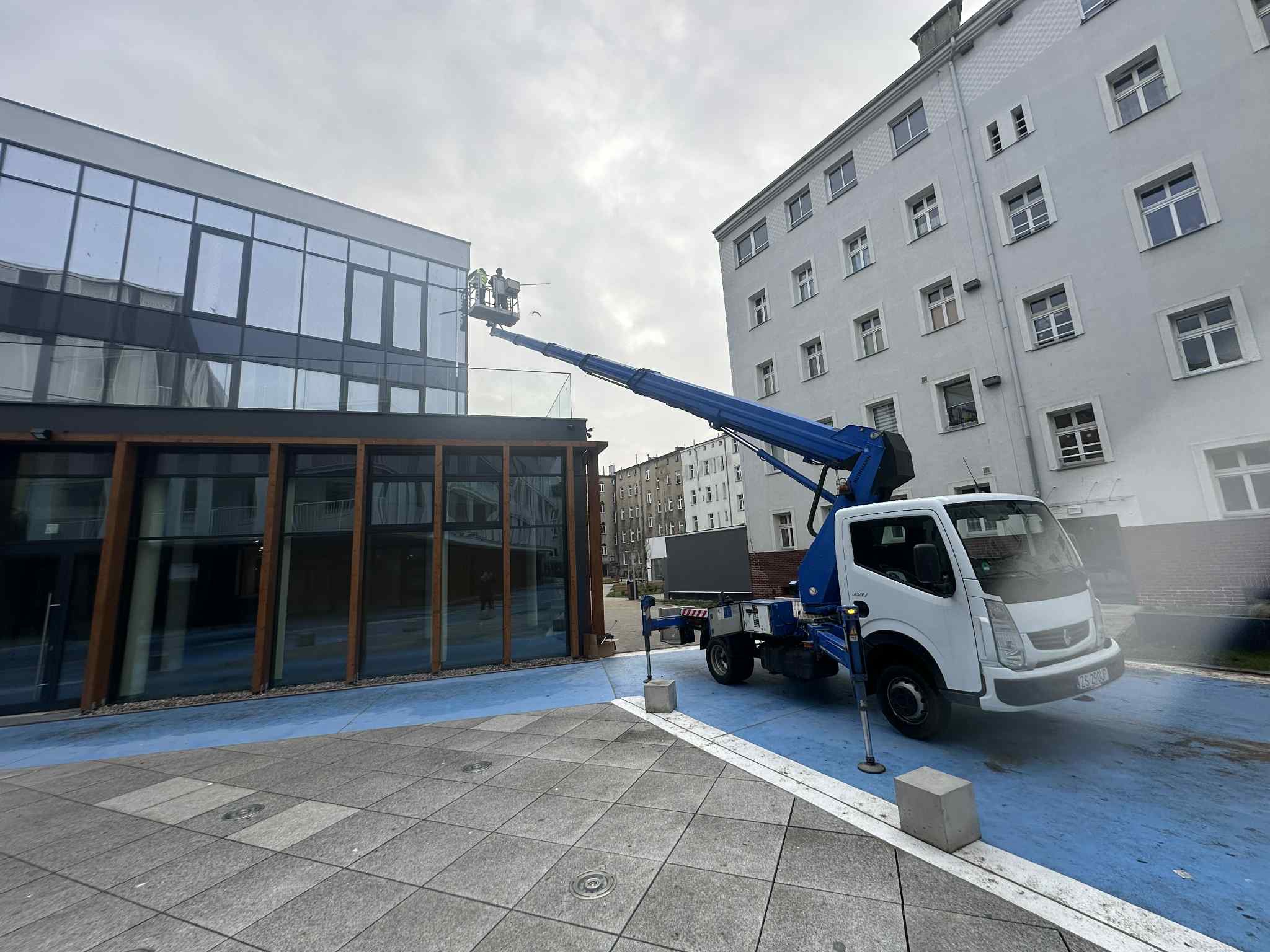 Pracownicy na podnośniku koszowym myją szklaną fasadę nowoczesnego budynku biurowego w pochmurny dzień. Widoczny fragment chodnika z szarej kostki.
