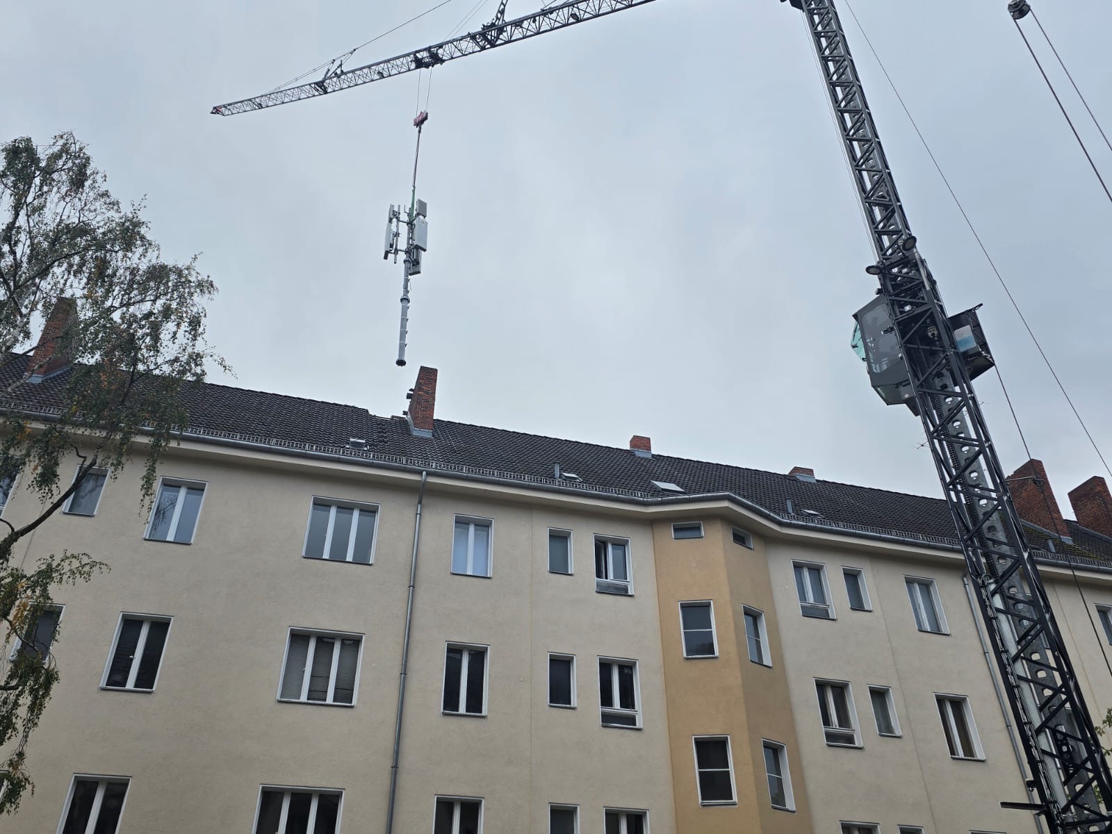 Montaż anteny telekomunikacyjnej na dachu budynku mieszkalnego za pomocą dźwigu. Widok z dołu na szary dach i elewację budynku.