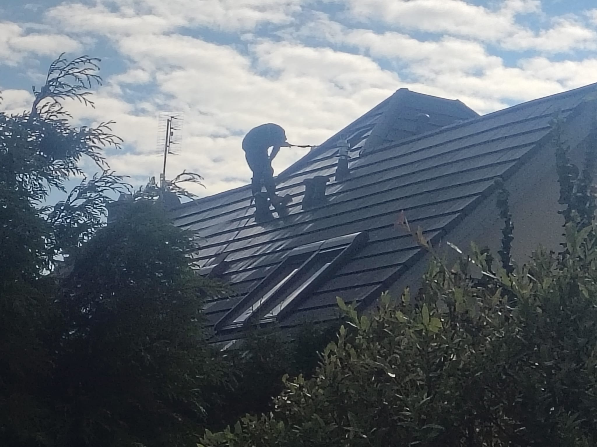 Praca na dachu domu z ciemną dachówką w słoneczny dzień. Widoczne okna dachowe i antena. Prace dekarskie w toku, sylwetka pracownika na tle nieba.