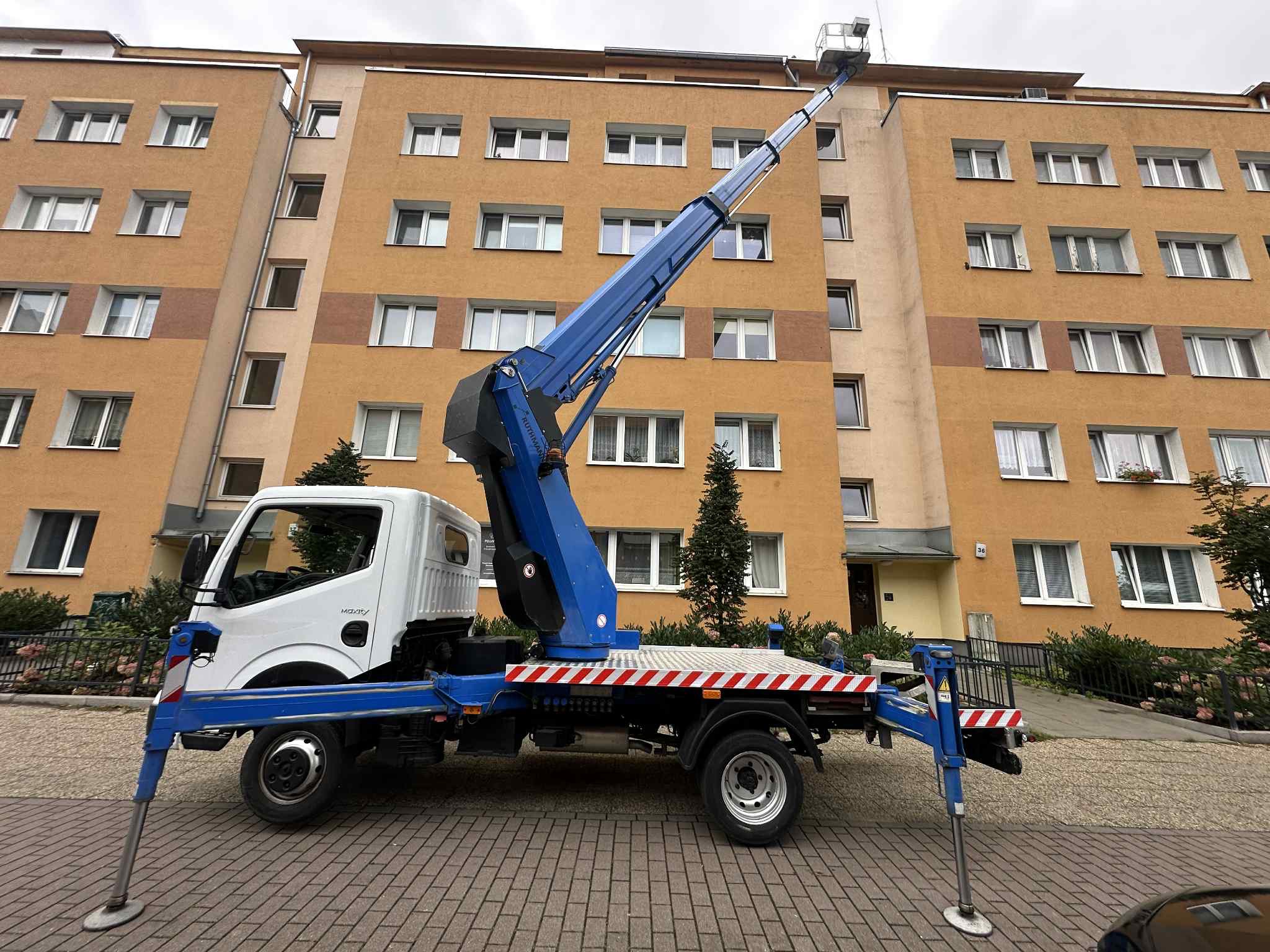Podnośnik koszowy na samochodzie na tle bloku mieszkalnego, wysięgnik skierowany ku górze, sugerujący prace na wysokościach.