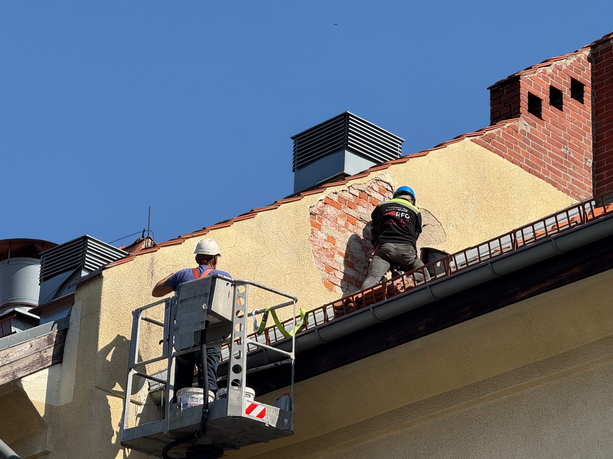 Dwóch pracowników na podnośniku naprawia uszkodzony fragment elewacji budynku z ceglanym kominem i metalowymi wylotami wentylacyjnymi na dachu.