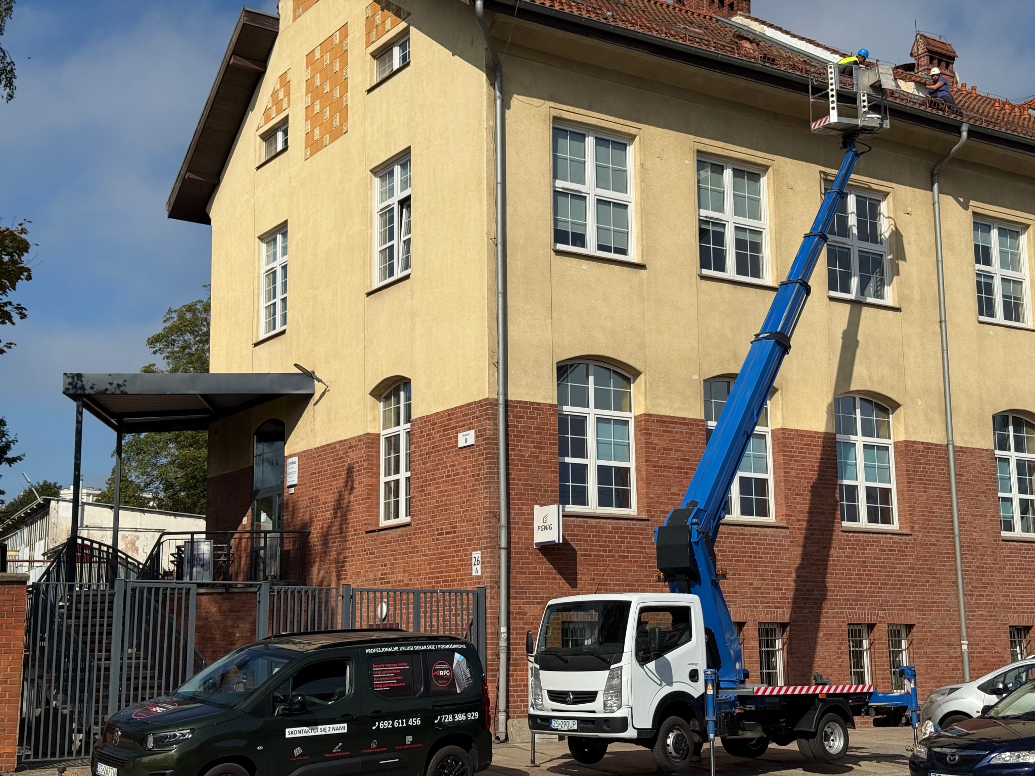 Praca na wysokości: podnośnik koszowy przy budynku z ceglaną fasadą i czerwonym dachem. Dwóch pracowników na platformie, prawdopodobnie podczas inspekcji lub naprawy dachu.