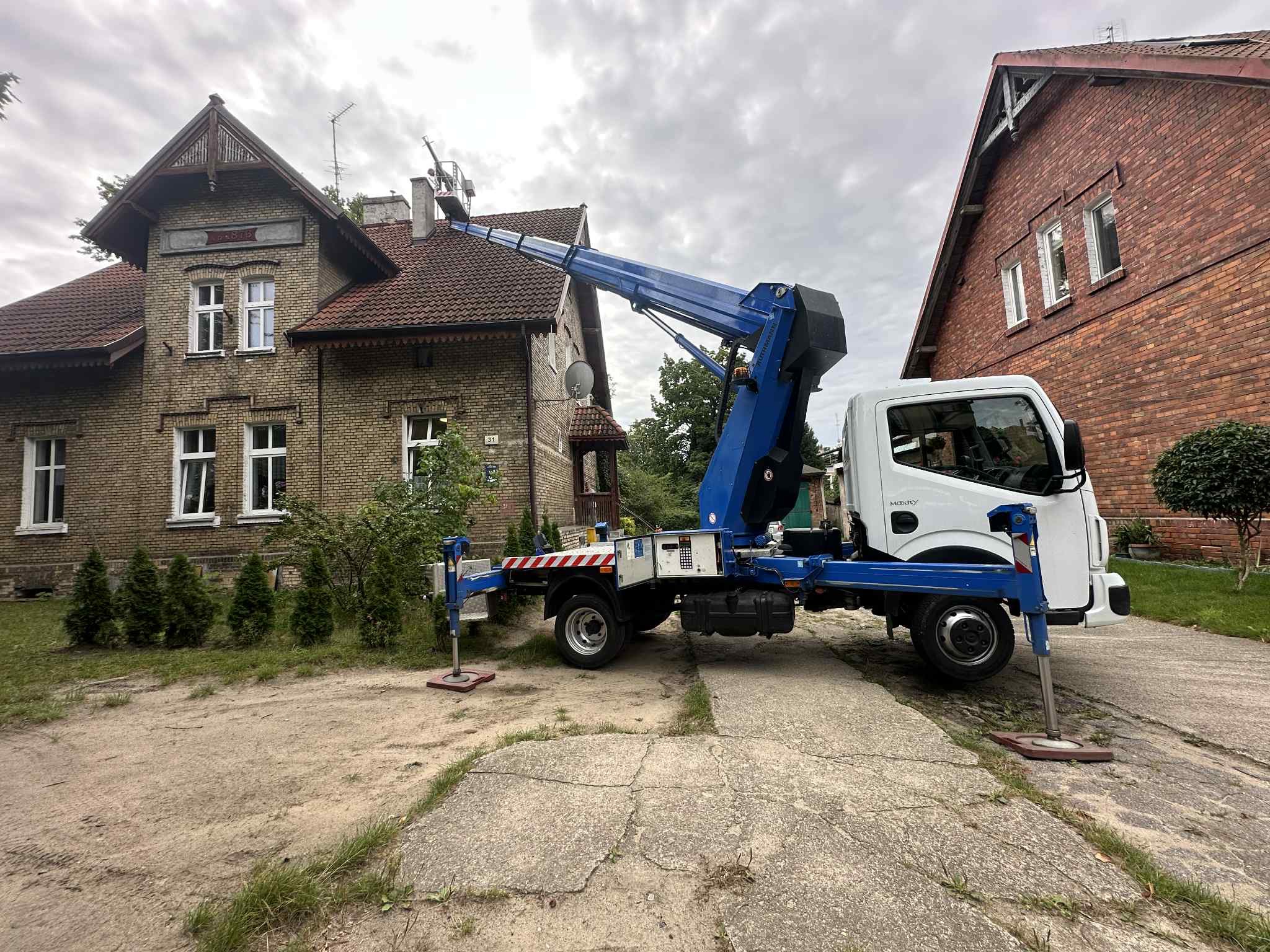 Podnośnik koszowy na tle ceglanego domu z czerwoną dachówką. Platforma robocza na wysokości dachu. Widoczne podpory stabilizujące pojazd.