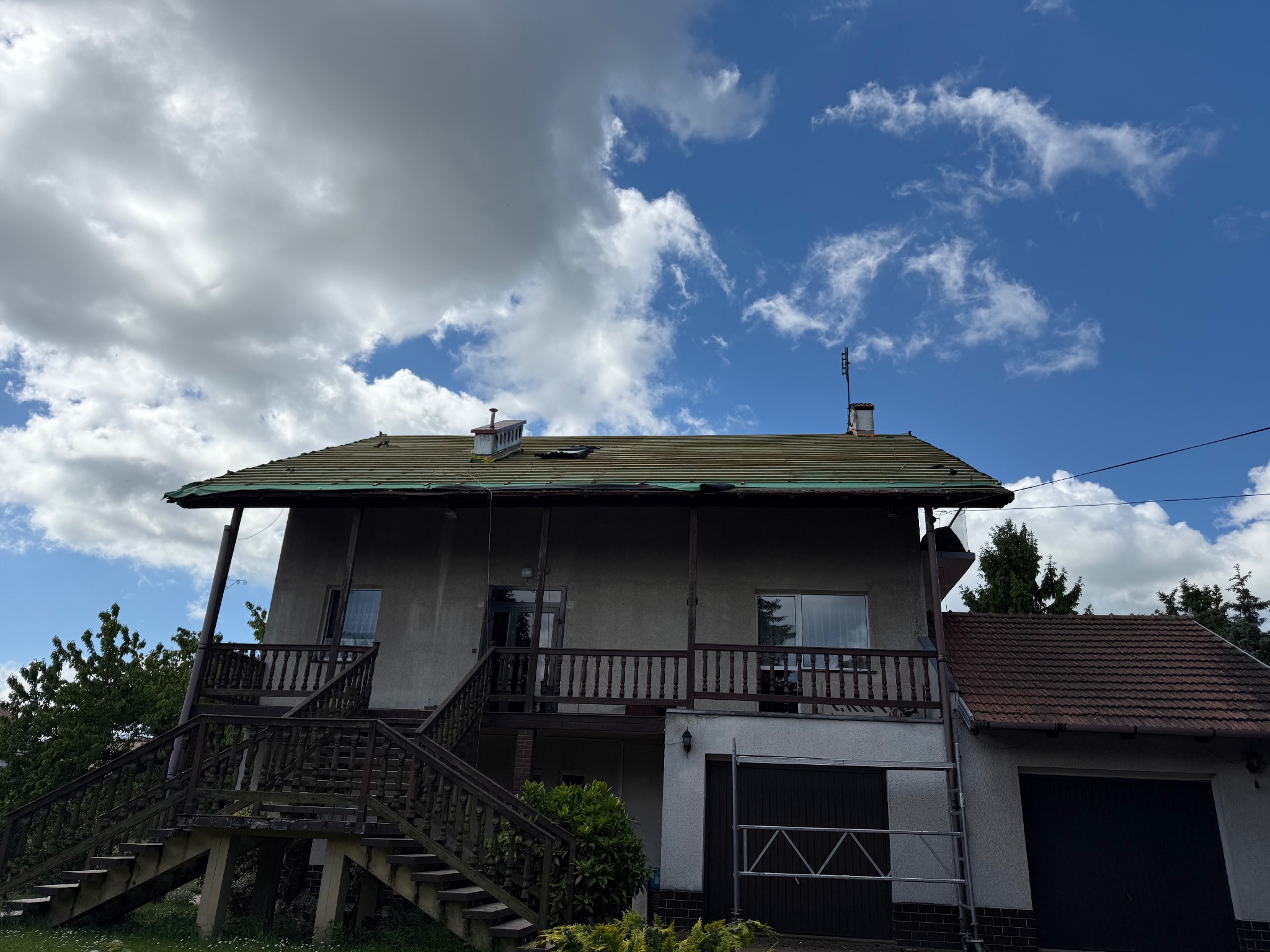 Dwukondygnacyjny dom z balkonem i zielonym dachem, widziany z dołu na tle błękitnego nieba z chmurami. Widoczne schody i garaż.