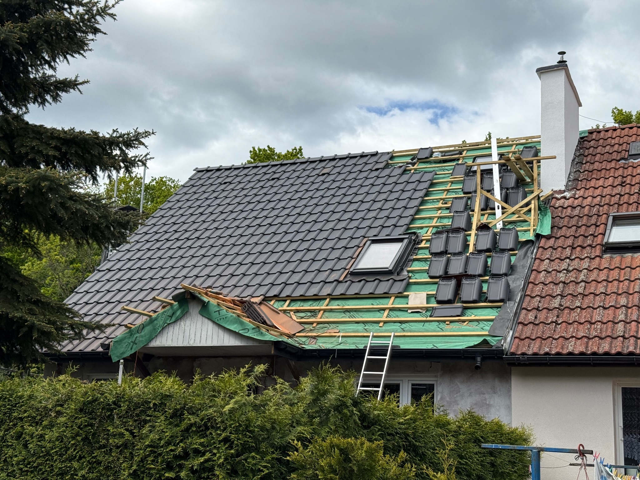 Częściowo pokryty dach z czarną dachówką, widoczne łaty i membrana. Na dachu drabina, obok komin i fragment dachu sąsiedniego budynku z czerwoną dachówką.
