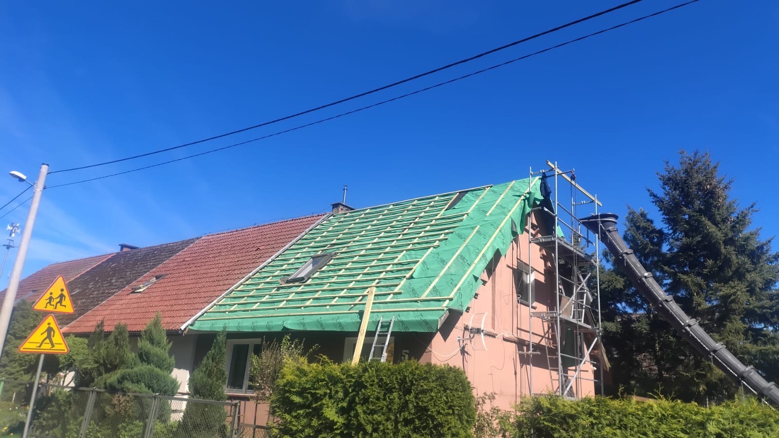 Dom w trakcie wymiany dachu: widoczne łaty, membrana dachowa, rusztowanie i zsyp na odpady. Część dachu z czerwoną dachówką pozostawiona. Słoneczny dzień.