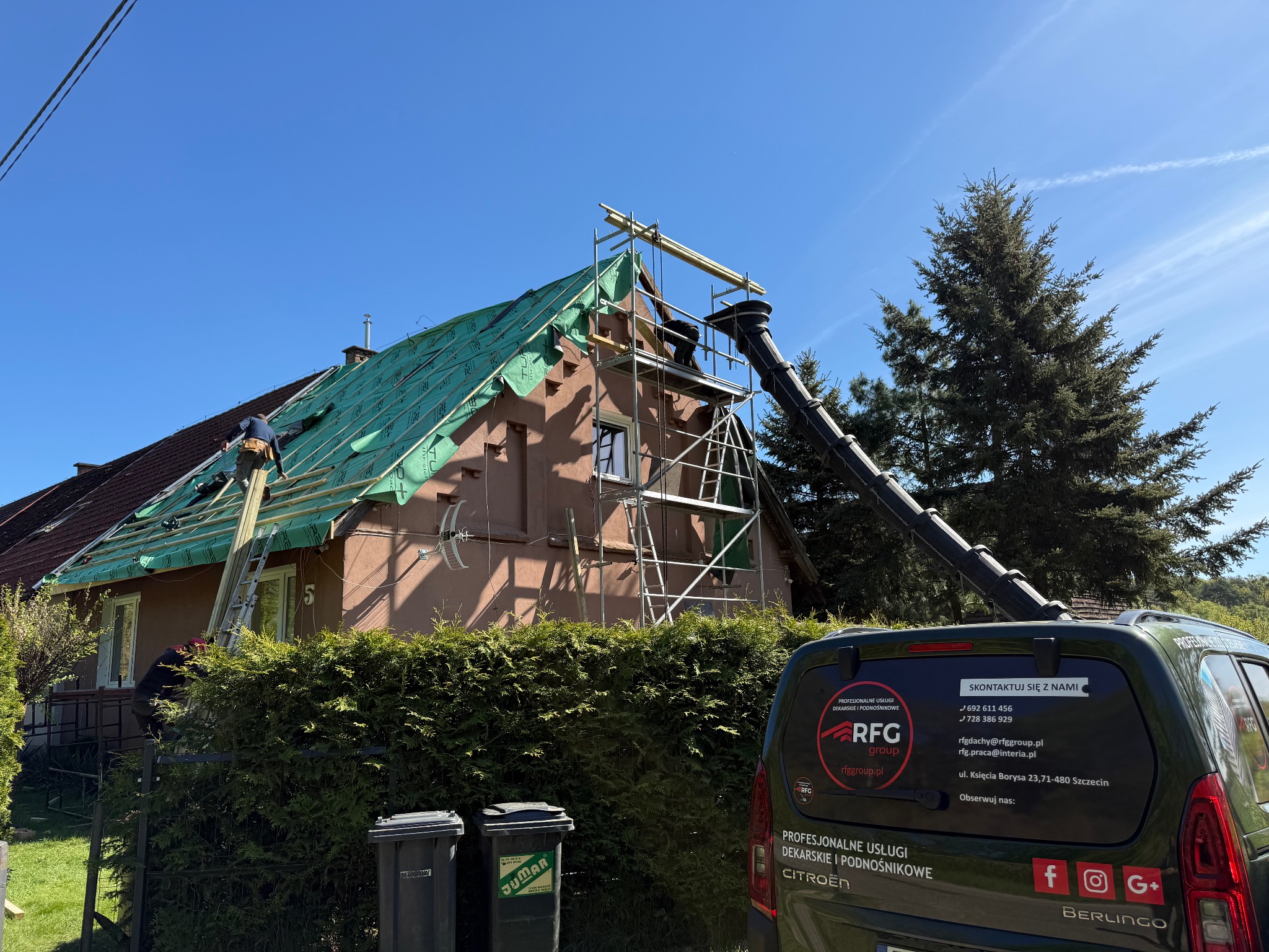 Dom w trakcie krycia dachu, widoczne rusztowania, ekipa dekarzy i zsyp gruzu. Na pierwszym planie samochód firmy dekarskiej.