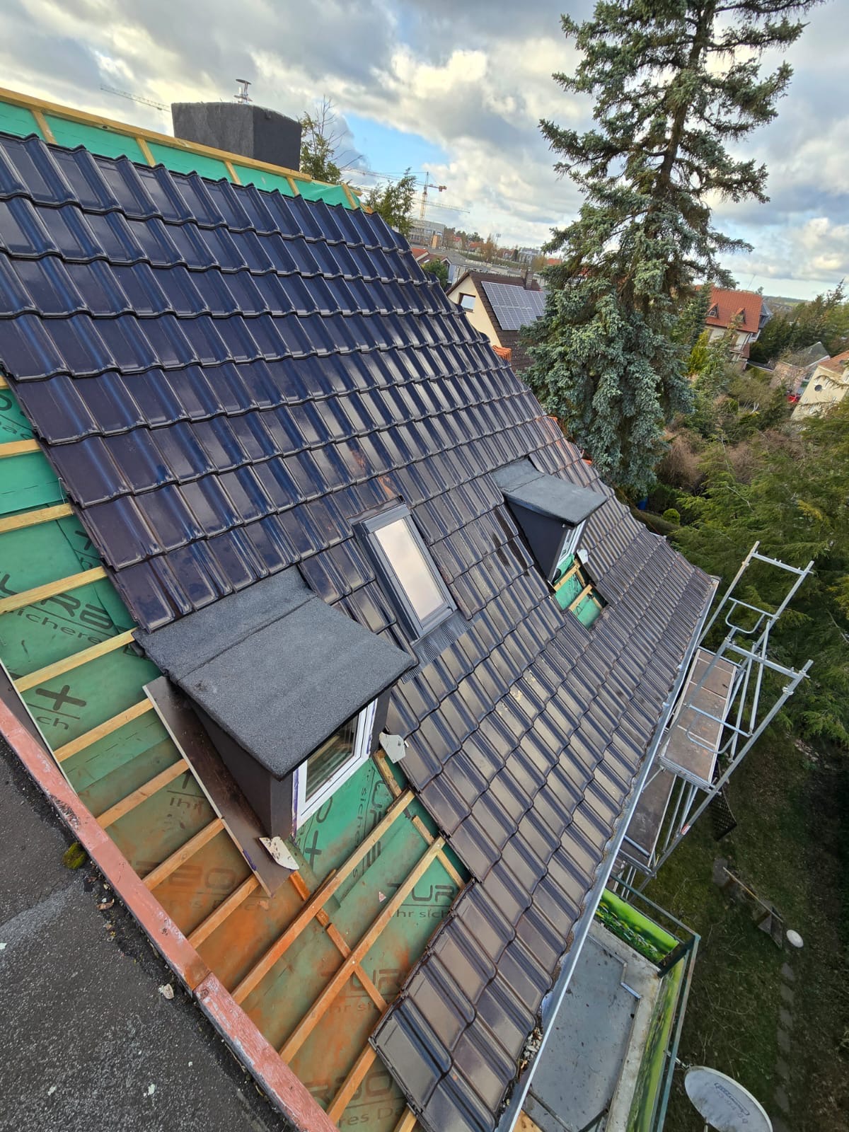 Częściowo pokryty dach z ciemnej dachówki ceramicznej, widoczne okna dachowe i fragment rusztowania. W tle drzewa i zabudowa mieszkalna.