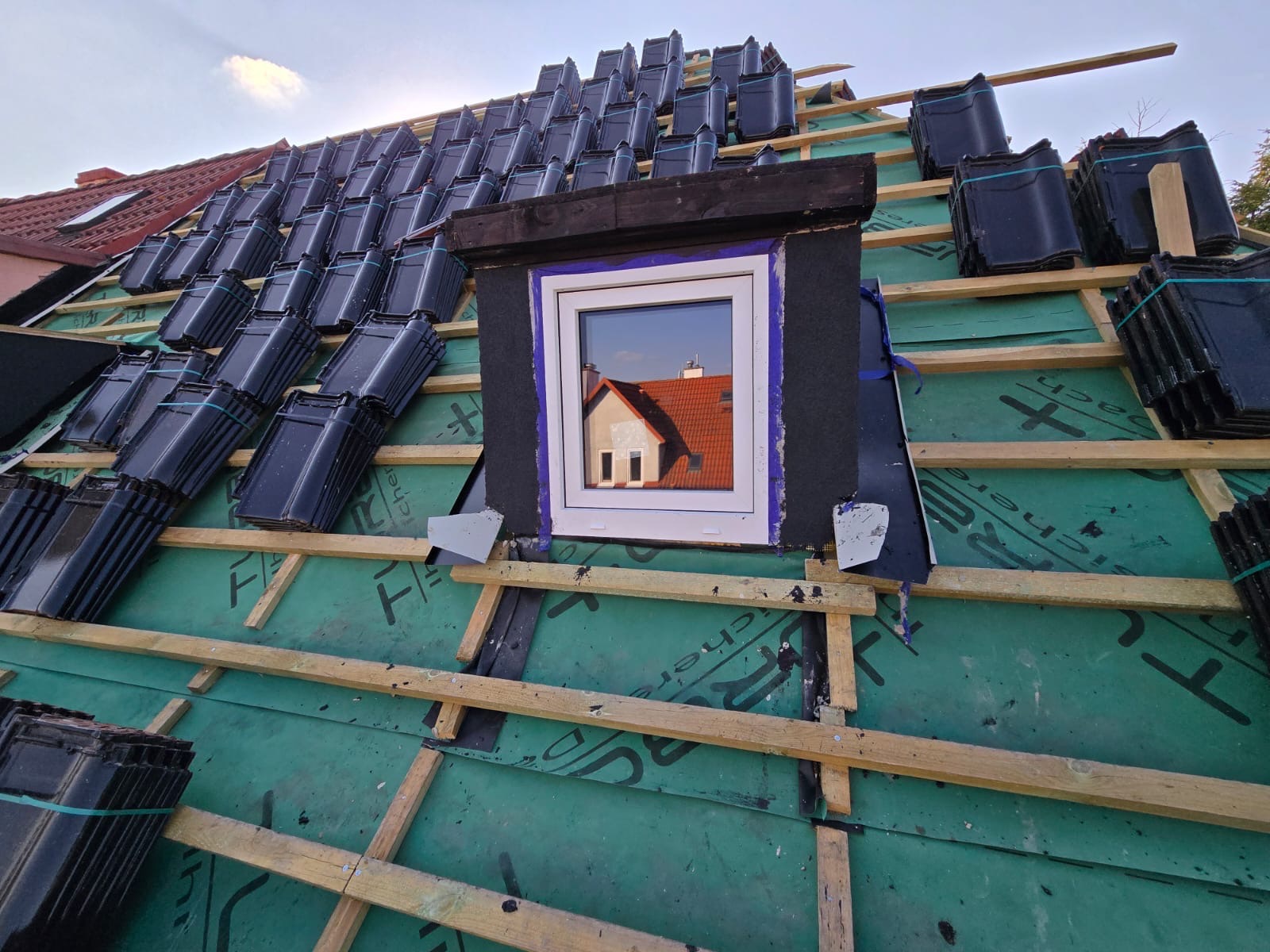 Dach w trakcie krycia dachówką ceramiczną, widoczne okno dachowe z odbiciem budynku w szybie, zielona membrana dachowa i drewniana konstrukcja.