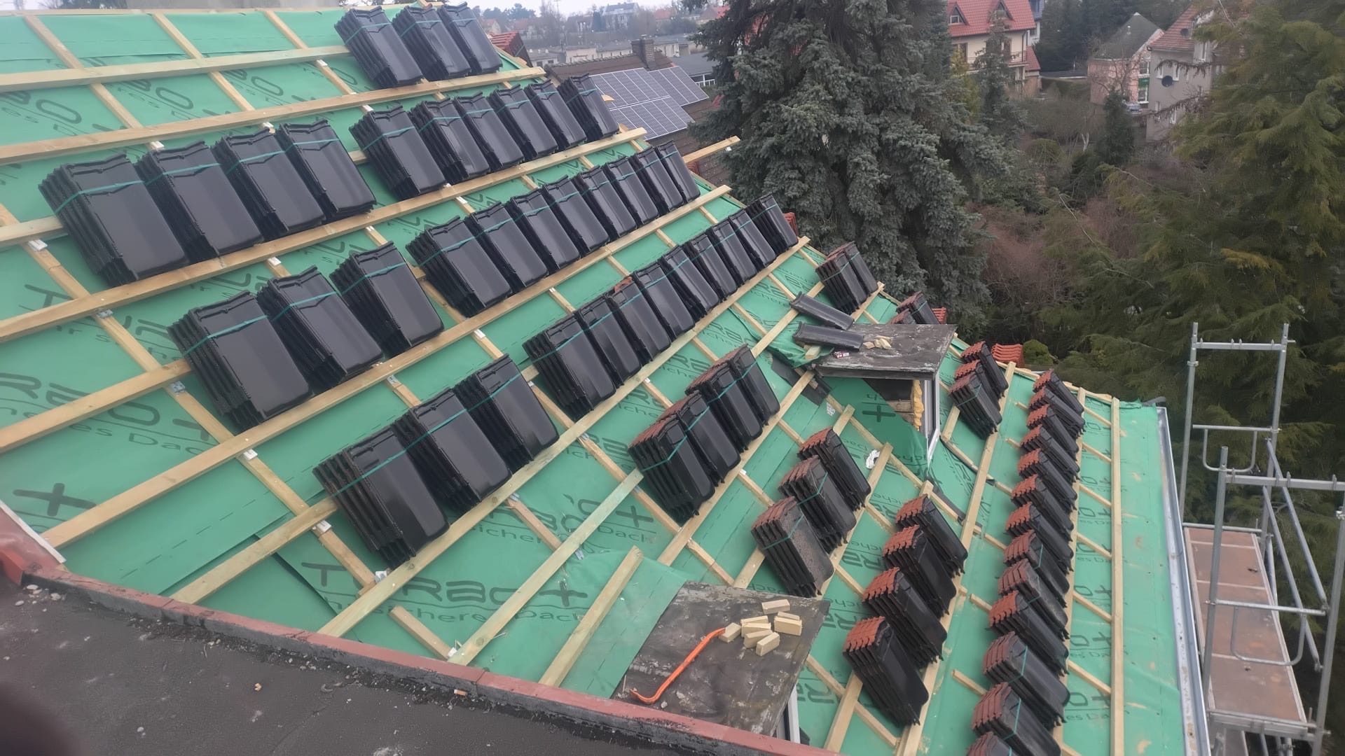 Układanie czarnej dachówki ceramicznej na zielonej membranie dachowej, widoczne drewniane łaty i narzędzia. Częściowo pokryty dach z widokiem na zabudowę.