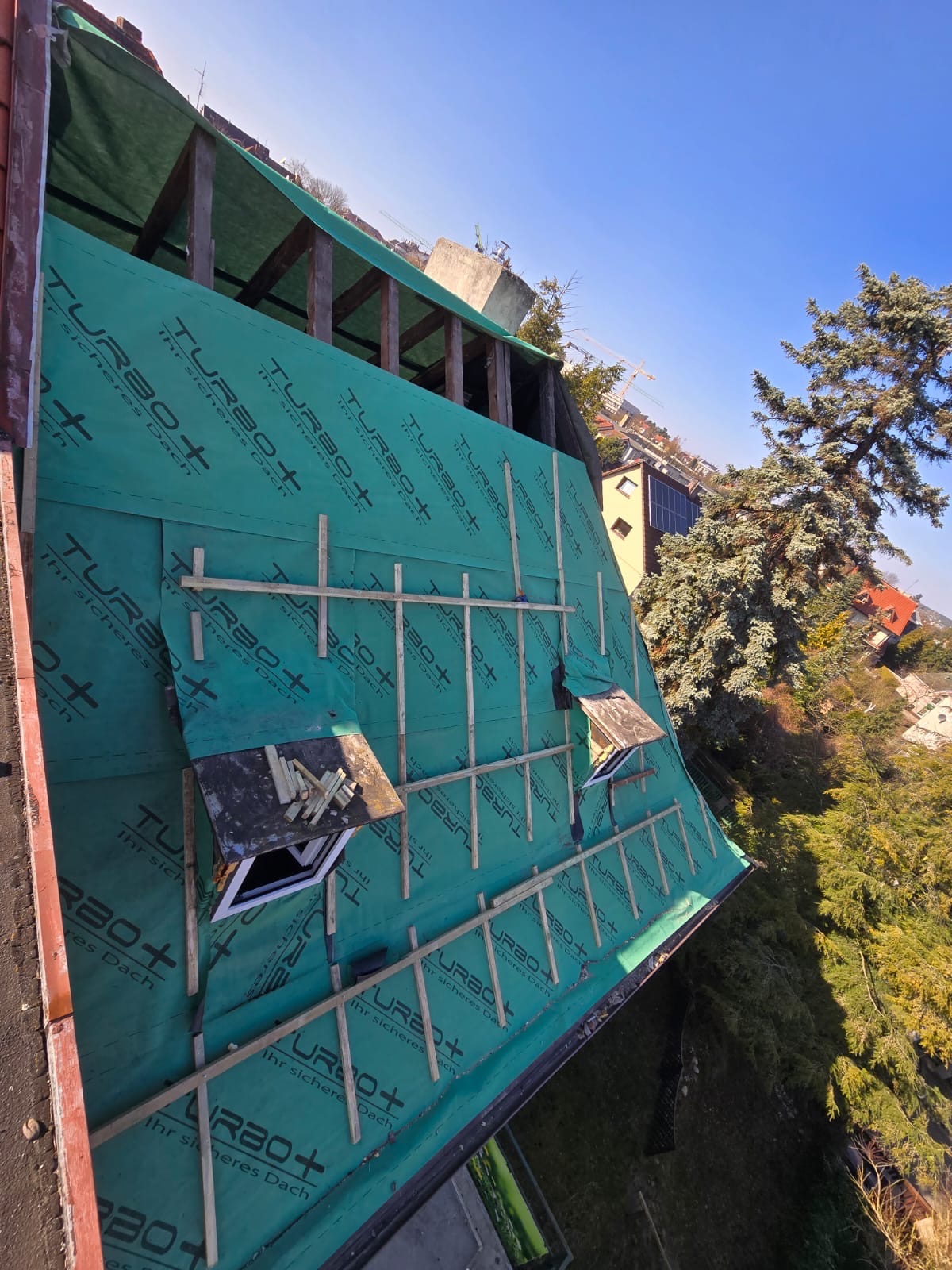 Dach w trakcie krycia, widoczna membrana TURBO+ i drewniana konstrukcja łat. Fragment okna dachowego z deskami. Ujęcie z góry, w tle zieleń i zabudowa miejska.