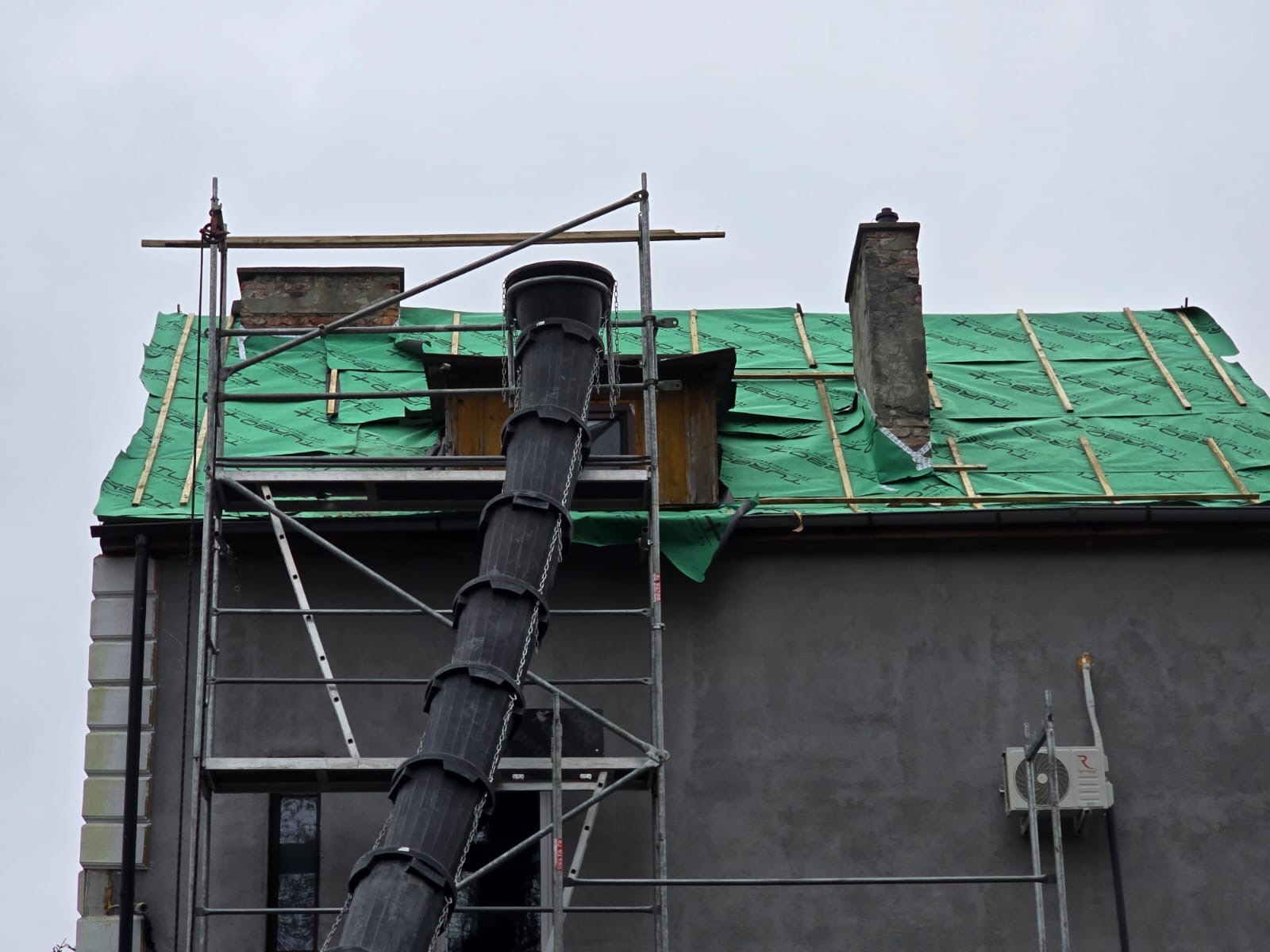 Dach z zieloną membraną, rusztowaniem i zsypem gruzu. Widoczny komin i okno dachowe. Prace dekarskie w toku, przygotowanie do krycia dachu.