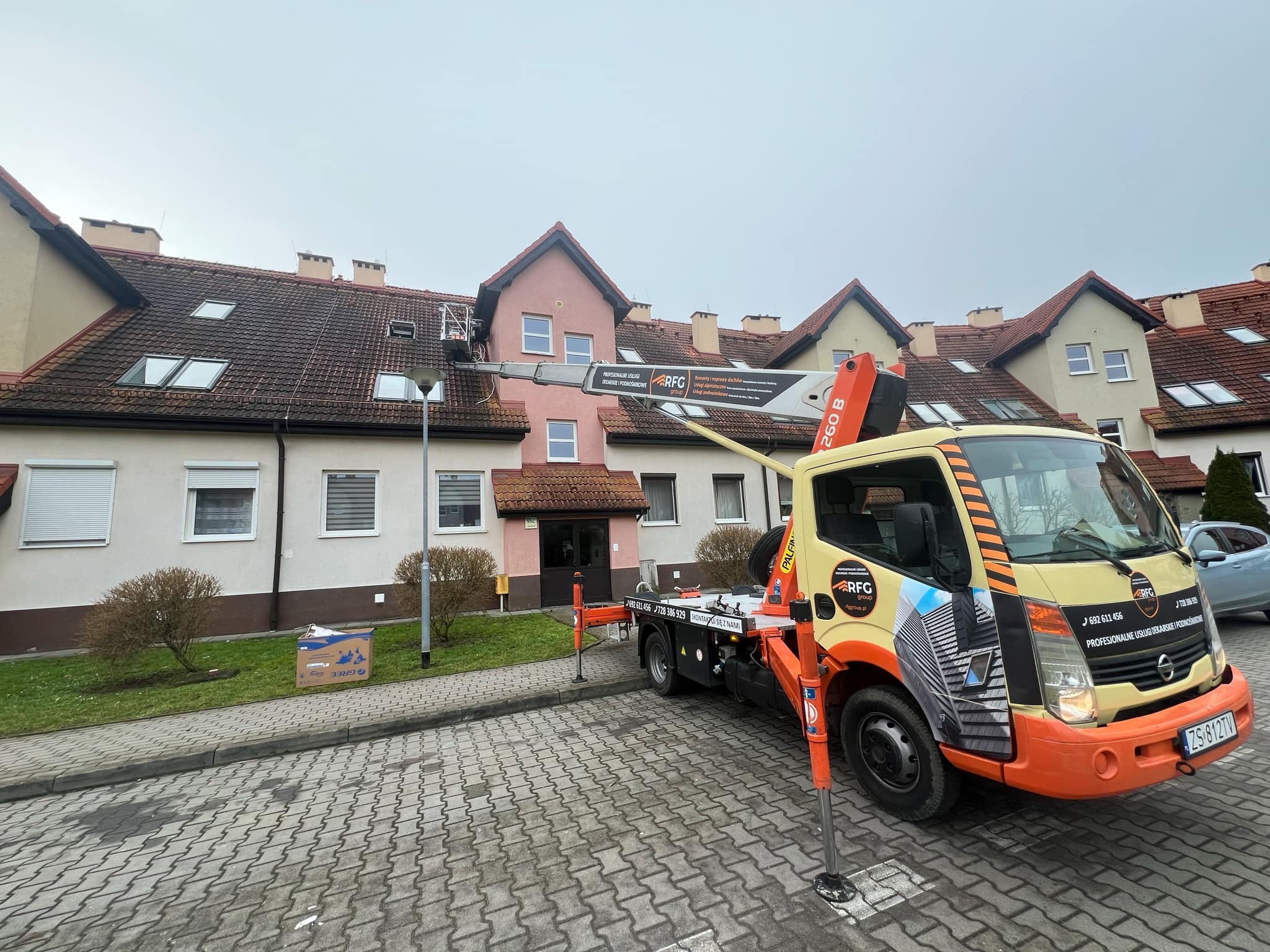 Podnośnik koszowy przy budynku mieszkalnym, prace dekarskie na dachu. Widoczny fragment dachu z oknami dachowymi i kominami. Żółto-pomarańczowy pojazd z logo firmy.
