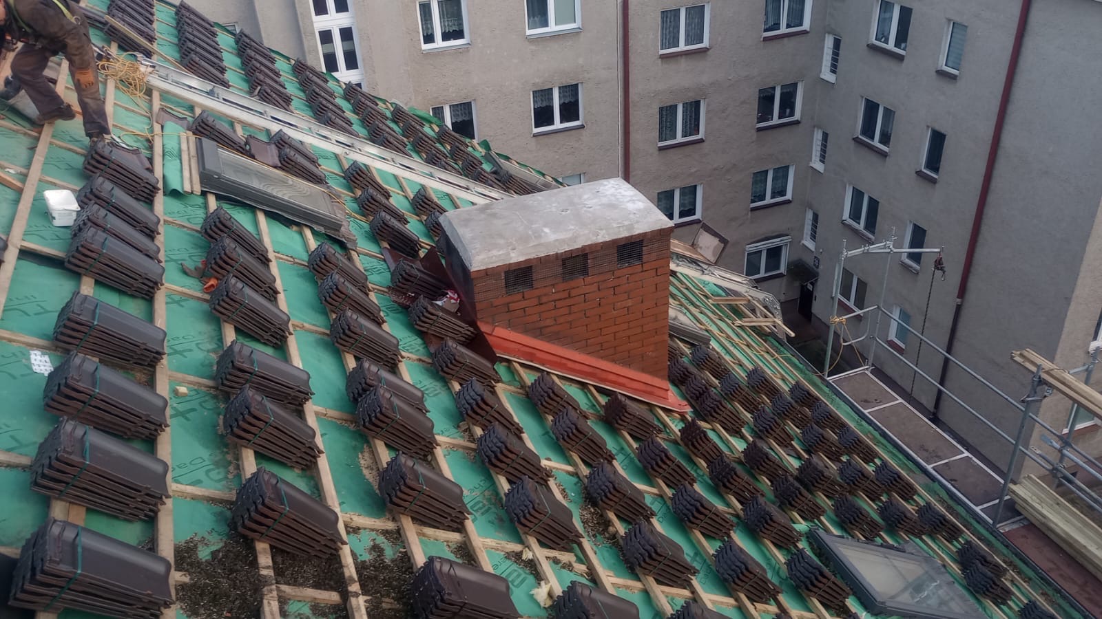 Układanie brązowej dachówki na zielonej membranie dachu budynku wielorodzinnego, widoczny komin i fragment rusztowania.