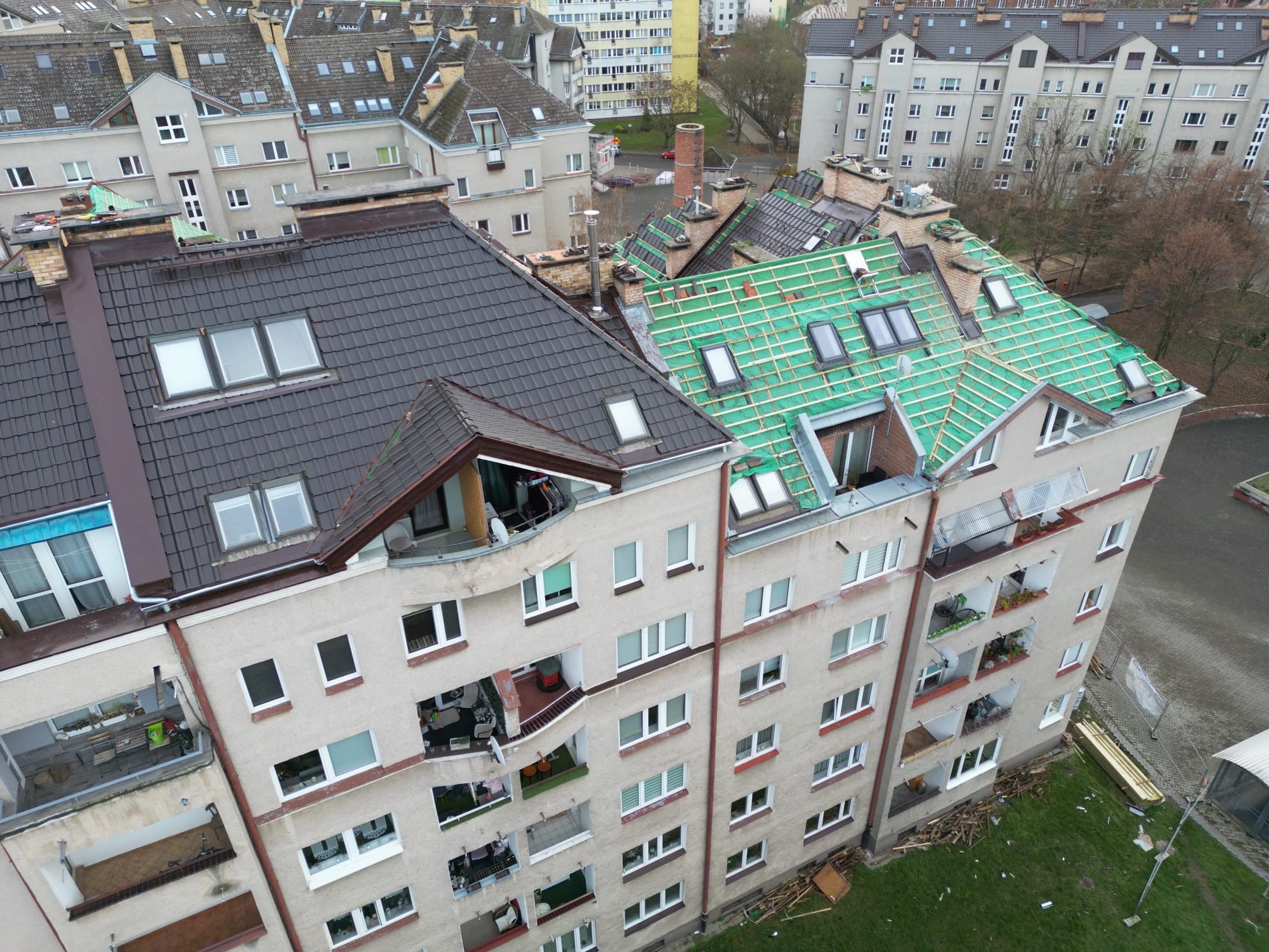 Wielorodzinny budynek z częściowo wymienionym dachem: ciemna dachówka po lewej, zielona membrana i łaty pod dachówkę po prawej. Widoczne okna dachowe i inne budynki w tle.
