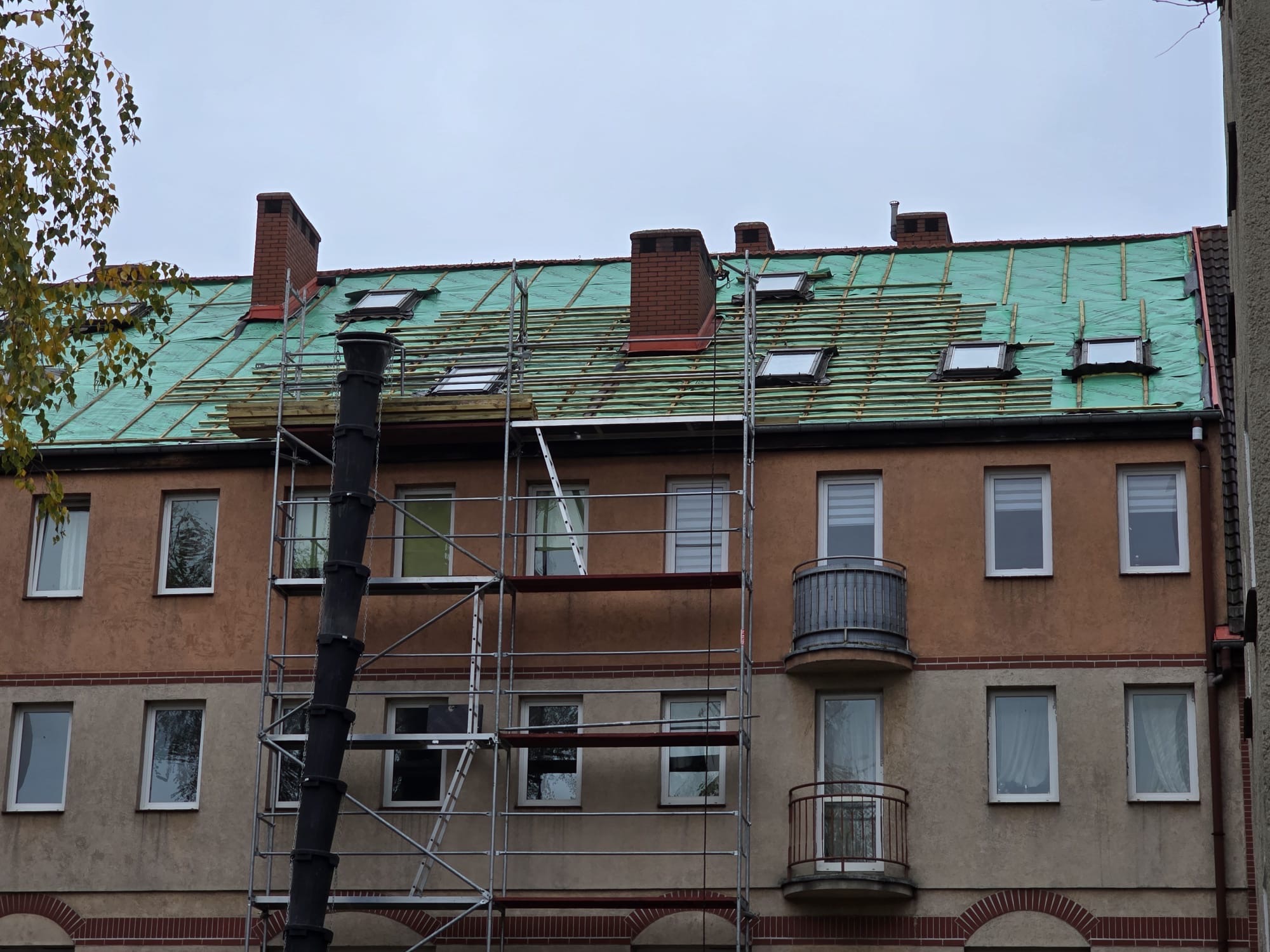 Budynek wielorodzinny z rusztowaniem i dachem w trakcie krycia, widoczne okna dachowe i kominy. Zabezpieczenie w kolorze zielonym pod konstrukcją dachu.