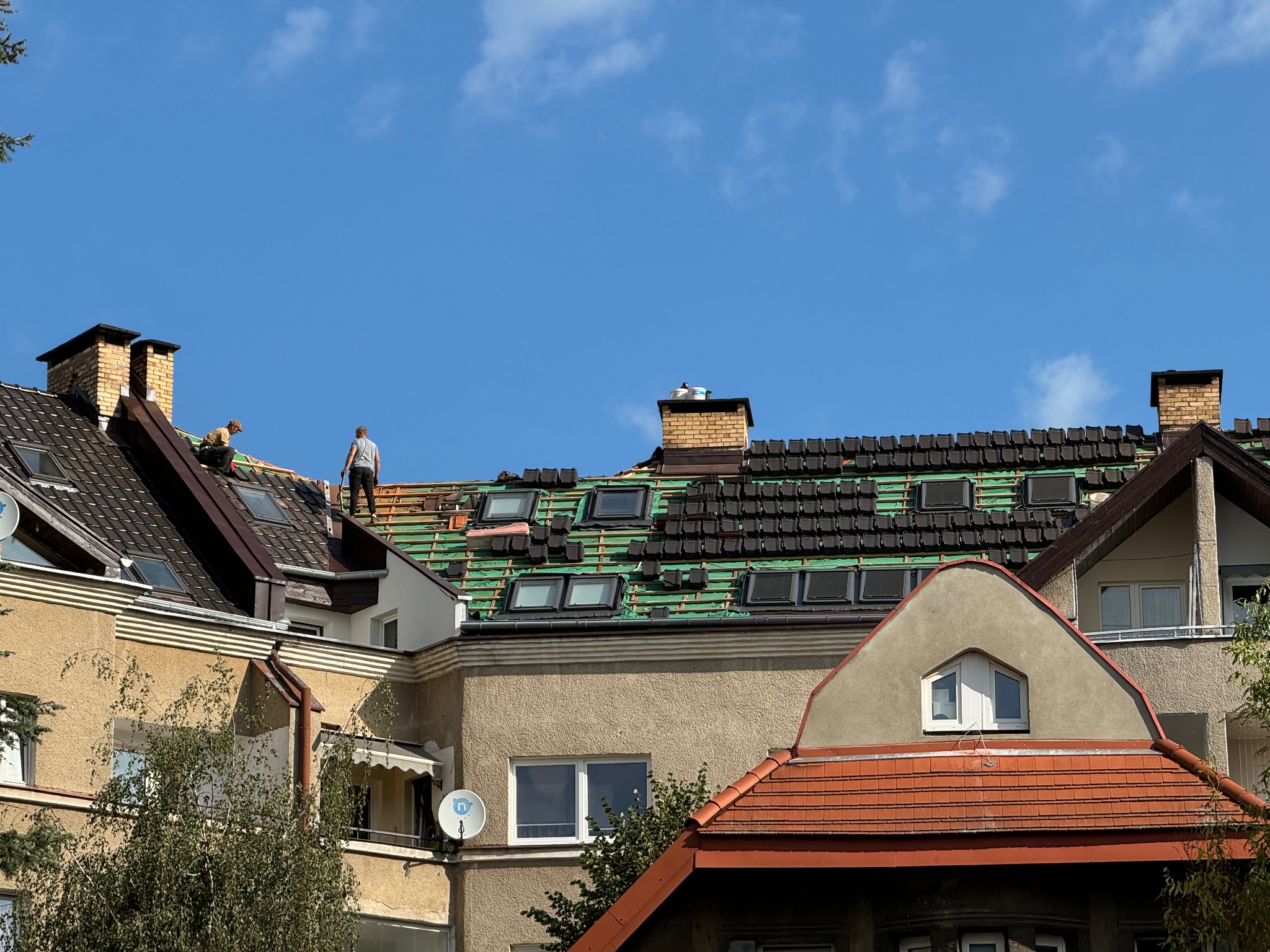 Fragment dachu z częściowo ułożoną dachówką, widoczne okna dachowe i dwóch dekarzy podczas pracy na tle błękitnego nieba z delikatnymi chmurami.