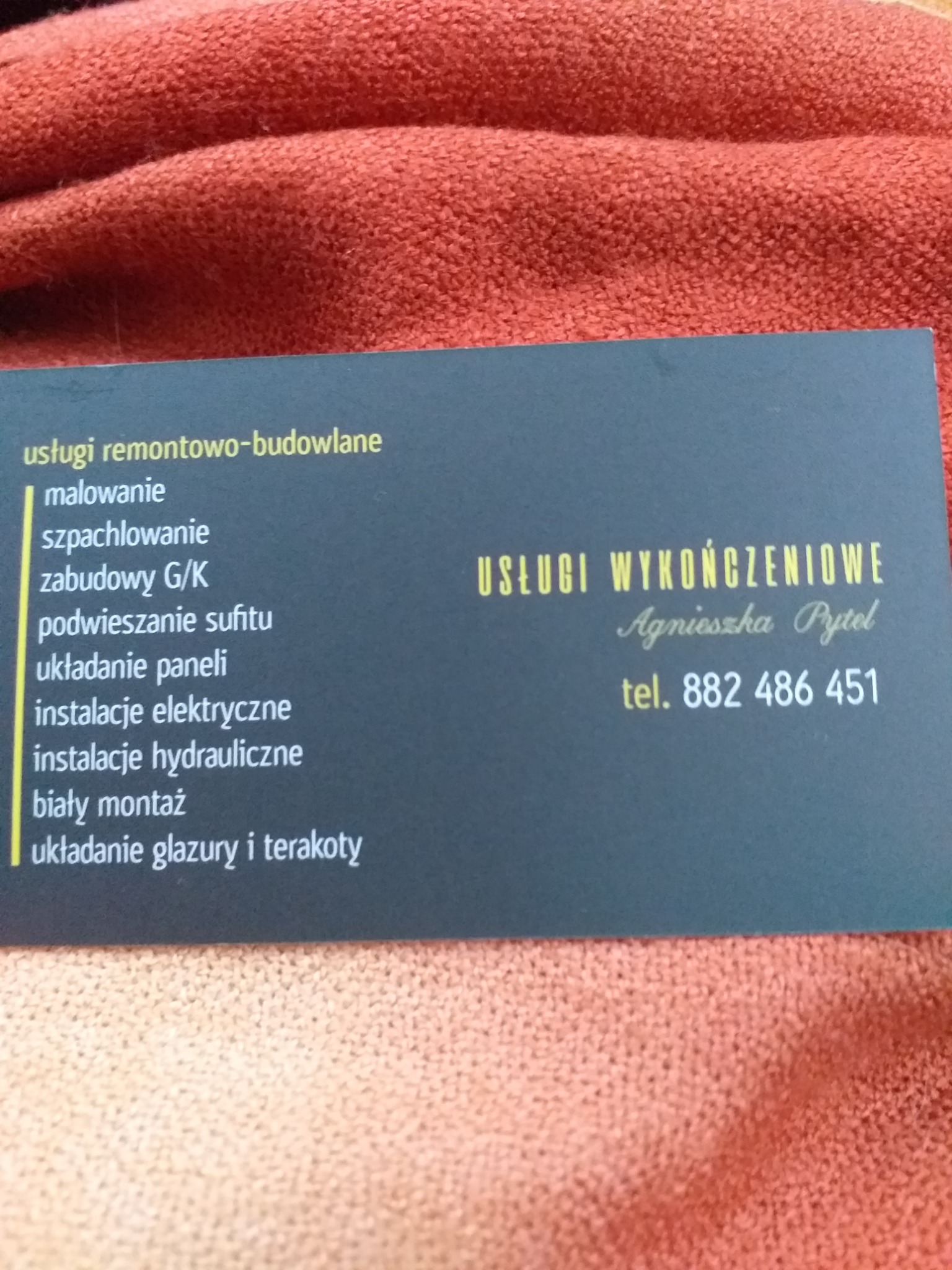 Wizytówka firmy remontowo-budowlanej 'Usługi Wykończeniowe Agnieszka Pytel' z numerem telefonu 882 486 451, leżąca na pomarańczowej tkaninie. Oferta obejmuje malowanie, szpachlowanie, zabudowy G/K,...