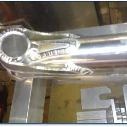 Precyzyjne spawy na metalowej ramie, widoczne regularne ściegi spawalnicze wokół okrągłego otworu i wzdłuż rury, z elementami mocującymi w tle.