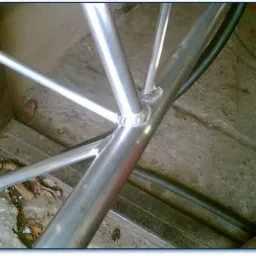 Spoina spawalnicza na konstrukcji z rur aluminiowych, widoczne połączenie kilku elementów pod kątem, surowe otoczenie warsztatowe.