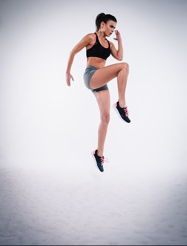 Kobieta w sportowym stroju wykonuje dynamiczne ćwiczenie unoszenia kolana w górę na jasnym tle.