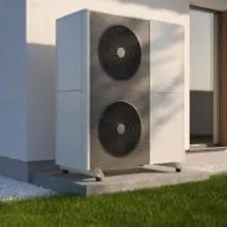 Zewnętrzna jednostka pompy ciepła typu powietrze-woda, umieszczona na betonowej podstawie przy ścianie budynku z dużymi oknami, otoczona trawnikiem i białym żwirem, oświetlona popołudniowym słońcem...