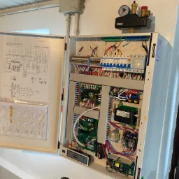 Otwarta szafka sterownicza z widocznymi modułami elektronicznymi, przewodami i schematem elektrycznym na drzwiach, zamontowana na białej ścianie.