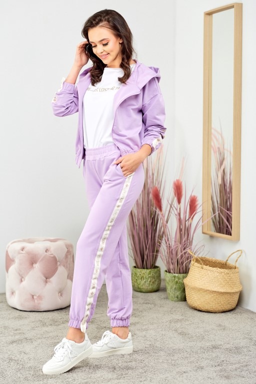 Kobieta w liliowym dresie z lampasami w Zduńskiej Woli, pozująca na tle lustra i dekoracyjnych traw w doniczkach.