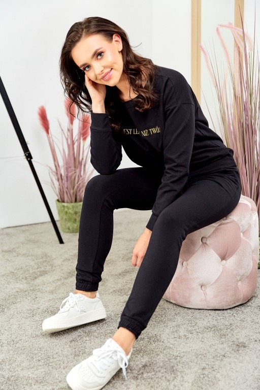 Kobieta siedzi na różowym pufie w czarnym dresie z napisem na bluzie i białych sneakersach, pozuje z ręką przy twarzy.